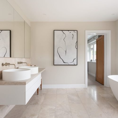 Bright, spacious bathroom featuring light travertine floor and wall tiles, a floating double-sink vanity, a large freestanding tub, and abstract line art.